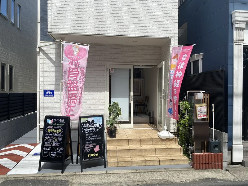 Tono Osteopathy の外観。白いタイルの階段の両脇に、産後骨盤矯正と自律神経ののぼりが2本立ち、入口の手前に黒板の立て看板が2つ置かれている