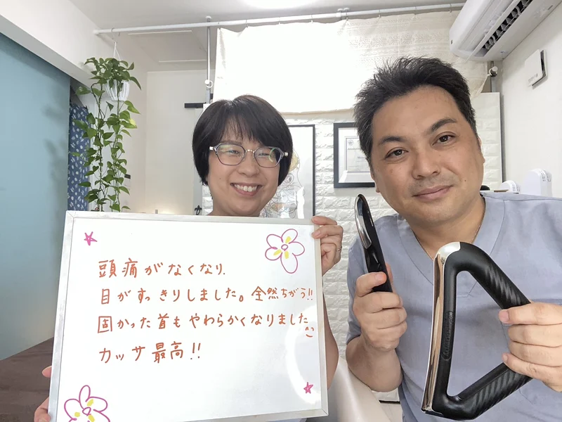 施術を受けた女性が「頭痛がなくなり、目がすっきりしました。全然ちがう!! 固かった首もやわらかくなりました。カッサ最高!!」と書いたボードを持ち、電動温熱式カッサを手にした院長と並んでいるところ